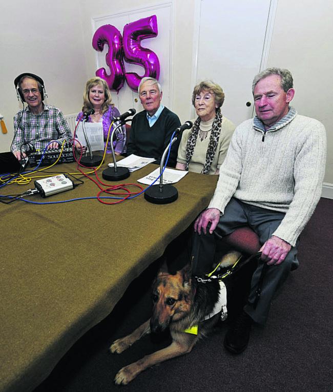 Three readers sit ready to record the news flanked on the left by a recording technician and on the right by a listener and his guide dog.
In the background a metallic purple helium balloon in the shape of a 35.
